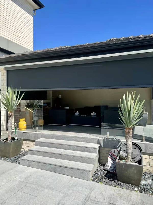 A dark outdoor blind shades a modern patio with glass railings and steps, offering privacy and sun protection for the entertainment area.
