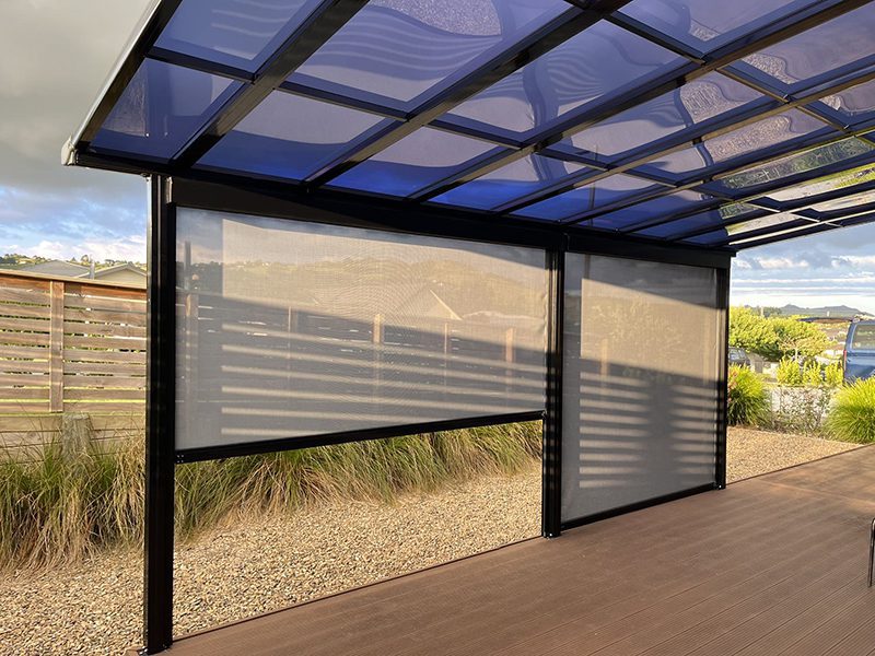 A set of light-colored windproof outdoor blinds are partially lowered on a patio, providing shade and protection under a clear-paneled pergola.